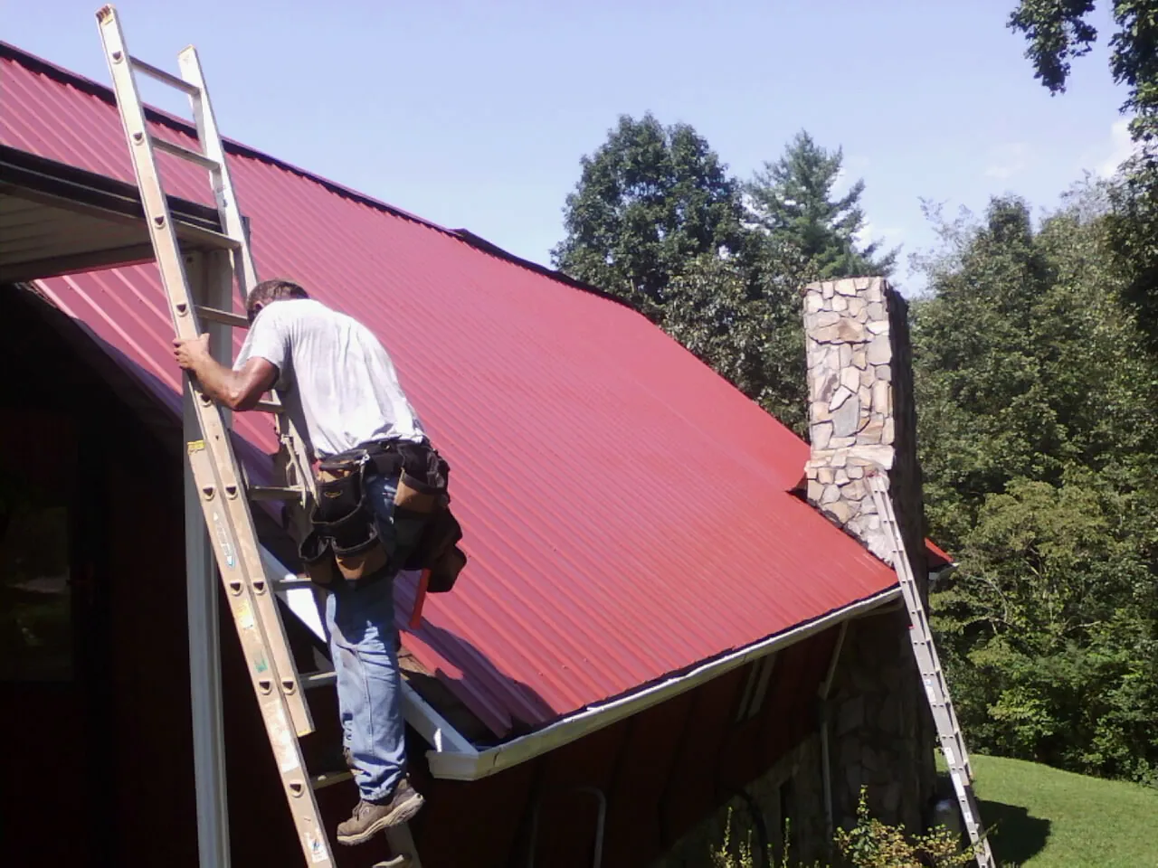 Roofer on a ladder beside a red metal roof and stone chimney — Soffit & Fascia Repair by Roofing Brothers in Amory